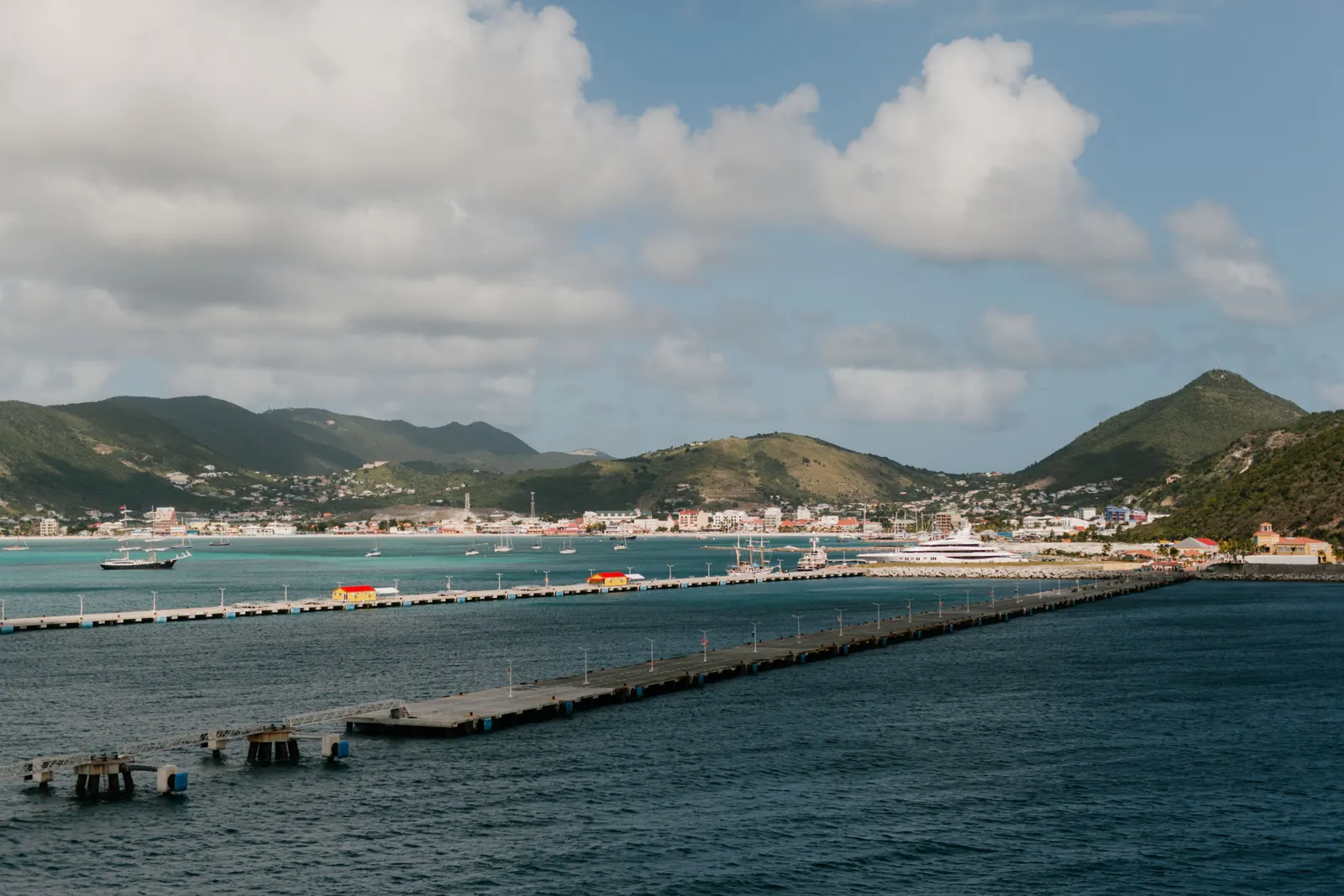 Tranquil harbor scene with piers, mountains, and vibrant town of St. Maarten under a bright sky.